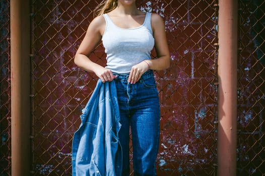 young woman in jeans clothes outdoors. portrait of a girl with freckles on her face, stylish girl on a background of a fence of a grid, on a sunny summer day. Holds a denim jacket in his hand