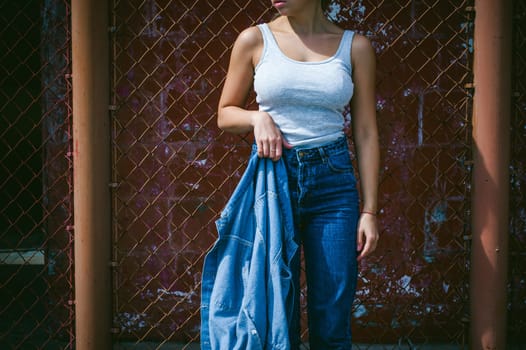 young woman in jeans clothes outdoors. portrait of a girl with freckles on her face, stylish girl on a background of a fence of a grid, on a sunny summer day. Holds a denim jacket in his hand