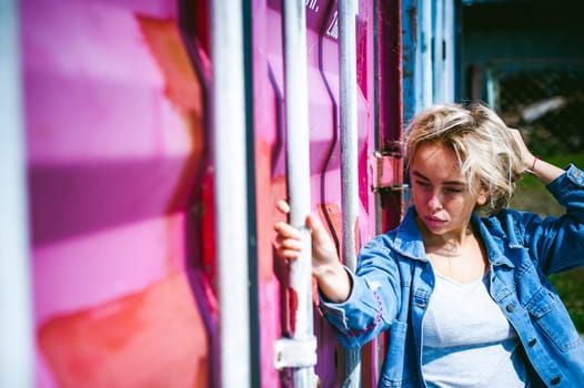young beautiful woman in jeans clothes outdoors. portrait of a girl with freckles on her face, stylish girl on a freight containers background, on a sunny summer day. wind develops her hair
