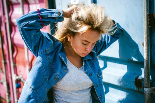 young beautiful woman in jeans clothes outdoors. portrait of a girl with freckles on her face, stylish girl on a freight containers background, on a sunny summer day. wind develops her hair