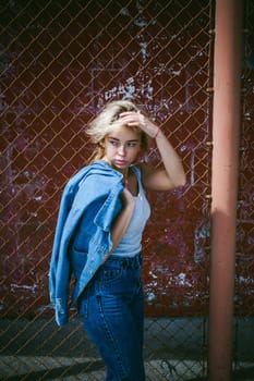 young woman in jeans clothes outdoors. portrait of a girl with freckles on her face, stylish girl on a background of a fence of a grid, on a sunny summer day. Holds a denim jacket in his hand