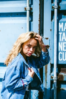 young beautiful woman in jeans clothes outdoors. portrait of a girl with freckles on her face, stylish girl on a freight containers background, on a sunny summer day. wind develops her hair