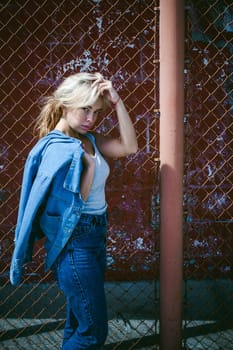 young woman in jeans clothes outdoors. portrait of a girl with freckles on her face, stylish girl on a background of a fence of a grid, on a sunny summer day. Holds a denim jacket in his hand