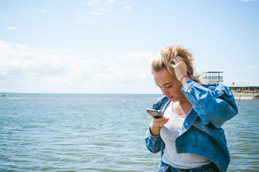 young beautiful woman in jeans clothes outdoors. portrait of a girl with freckles on her face, stylish girl on sea beach, on a sunny summer autumn day. looks at mobile phone