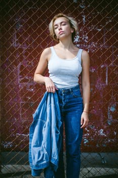 young woman in jeans clothes outdoors. portrait of a girl with freckles on her face, stylish girl on a background of a fence of a grid, on a sunny summer day. Holds a denim jacket in his hand