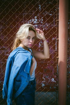young woman in jeans clothes outdoors. portrait of a girl with freckles on her face, stylish girl on a background of a fence of a grid, on a sunny summer day. Holds a denim jacket in his hand