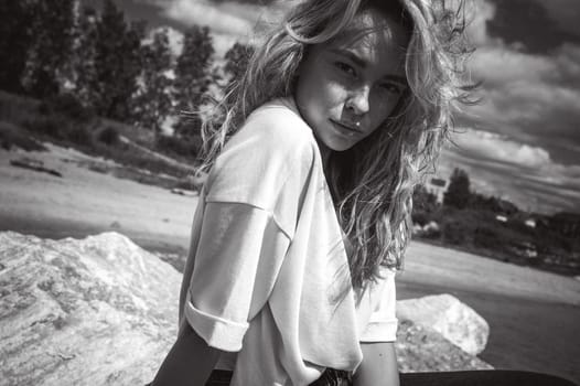 Portrait young woman on stones near sea. Beautiful girl is resting on coast, enjoying outdoor recreation.