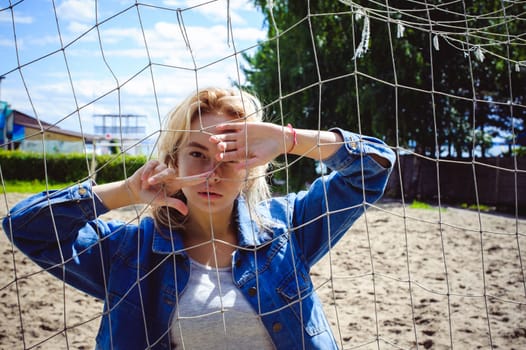 young woman in jeans clothes outdoors. portrait of a girl with freckles on her face, against the background of a football gate grid, on a sunny summer day. hands and fingers