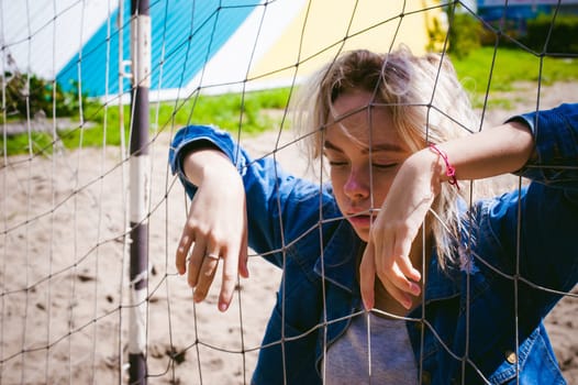 young woman in jeans clothes outdoors. portrait of a girl with freckles on her face, against the background of a football gate grid, on a sunny summer day. hands and fingers