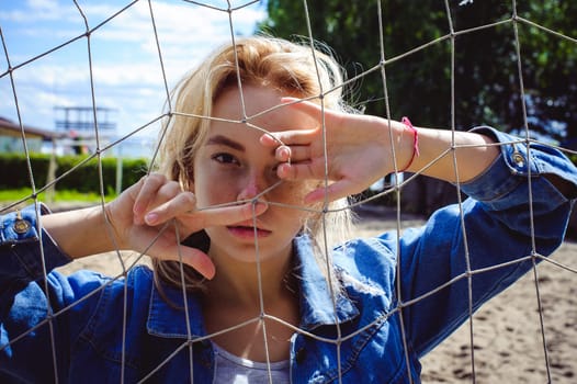 young woman in jeans clothes outdoors. portrait of a girl with freckles on her face, against the background of a football gate grid, on a sunny summer day. hands and fingers