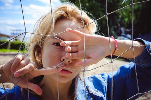 young woman in jeans clothes outdoors. portrait of a girl with freckles on her face, against the background of a football gate grid, on a sunny summer day. hands and fingers