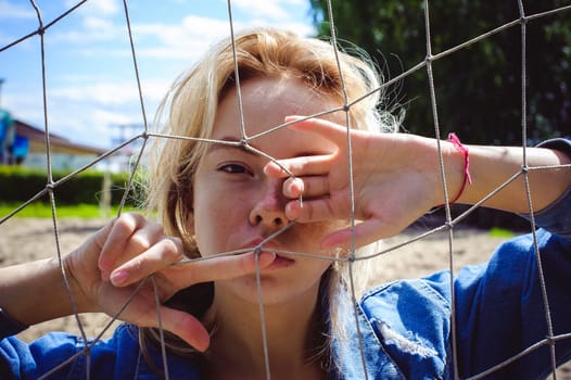 young woman in jeans clothes outdoors. portrait of a girl with freckles on her face, against the background of a football gate grid, on a sunny summer day. hands and fingers