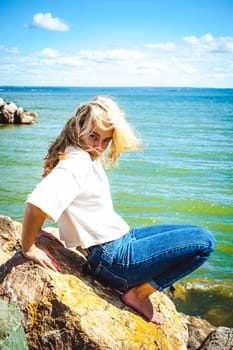 Portrait young woman on stones near sea. Beautiful girl is resting on coast, enjoying outdoor recreation.