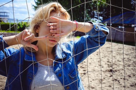 young woman in jeans clothes outdoors. portrait of a girl with freckles on her face, against the background of a football gate grid, on a sunny summer day. hands and fingers