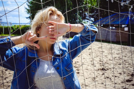 young woman in jeans clothes outdoors. portrait of a girl with freckles on her face, against the background of a football gate grid, on a sunny summer day. hands and fingers