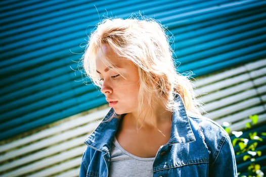 young woman in jeans clothes outdoors. portrait of a girl with freckles on her face, against the background of a metal striped fence, on a sunny summer day