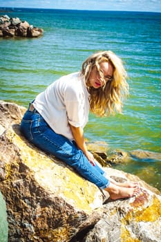 Portrait young woman on stones near sea. Beautiful girl is resting on coast, enjoying outdoor recreation.