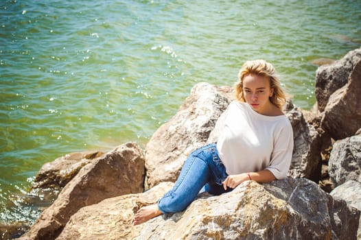 Portrait young woman on stones near sea. Beautiful girl is resting on coast, enjoying outdoor recreation.