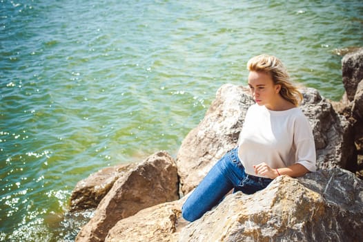 Portrait young woman on stones near sea. Beautiful girl is resting on coast, enjoying outdoor recreation.