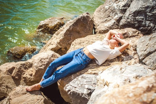 Portrait young woman on stones near sea. Beautiful girl is resting on coast, enjoying outdoor recreation.
