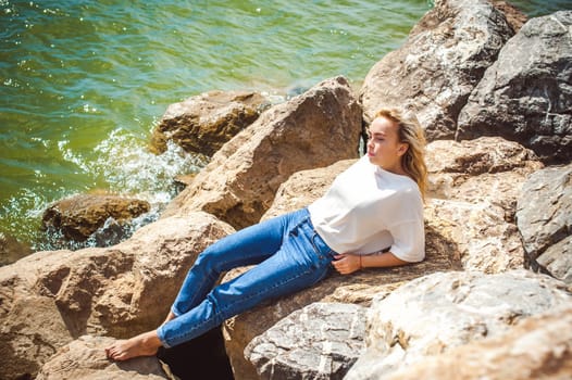 Portrait young woman on stones near sea. Beautiful girl is resting on coast, enjoying outdoor recreation.