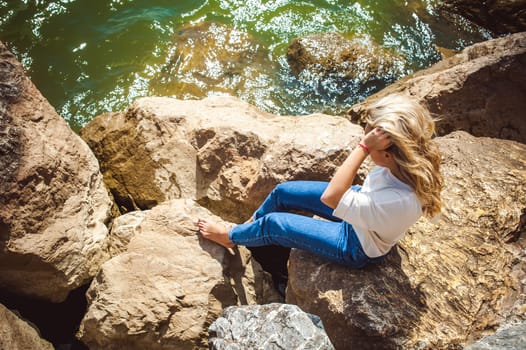 Portrait young woman on stones near sea. Beautiful girl is resting on coast, enjoying outdoor recreation.