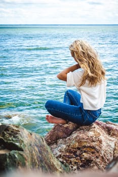 Portrait young woman on stones near sea. Beautiful girl is resting on coast, enjoying outdoor recreation.