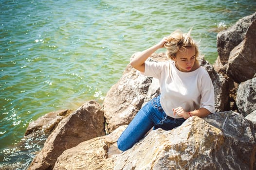 Portrait young woman on stones near sea. Beautiful girl is resting on coast, enjoying outdoor recreation.