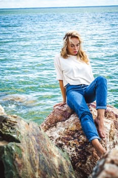 Portrait young woman on stones near sea. Beautiful girl is resting on coast, enjoying outdoor recreation.