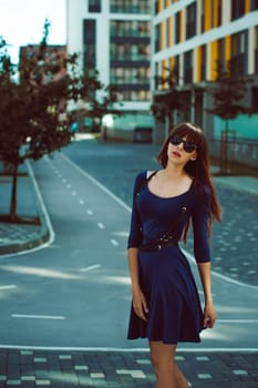 Young attractive woman with long legs with long hair in a blue elegant dress with a sword belt In sunglasses, strolls in a European modern courtyard, a warm summer day