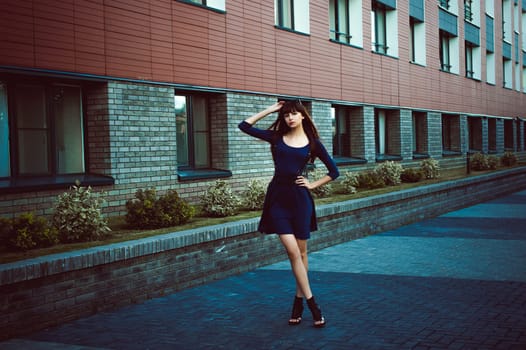 Young attractive woman with long legs with long hair in a blue elegant dress with a sword belt, strolls in a European modern courtyard, a warm summer day