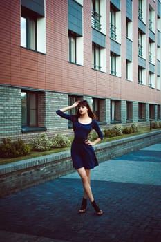 Young attractive woman with long legs with long hair in a blue elegant dress with a sword belt, strolls in a European modern courtyard, a warm summer day