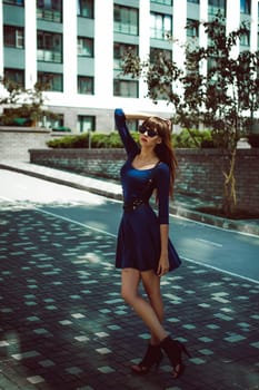 Young attractive woman with long legs with long hair in a blue elegant dress with a sword belt In sunglasses, strolls in a European modern courtyard, a warm summer day