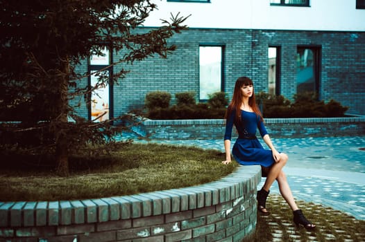 Young attractive woman with long legs with long hair in a blue elegant dress with a sword belt, strolls in a European modern courtyard, a warm summer day