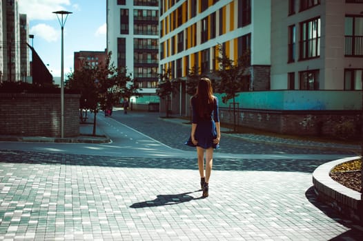 Young attractive woman with long legs with long hair in a blue elegant dress with a sword belt, strolls in a European modern courtyard, a warm summer day