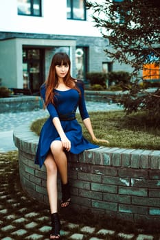 Young attractive woman with long legs with long hair in a blue elegant dress with a sword belt, strolls in a European modern courtyard, a warm summer day