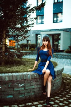 Young attractive woman with long legs with long hair in a blue elegant dress with a sword belt, strolls in a European modern courtyard, a warm summer day