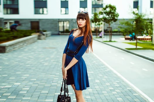 Young attractive woman with long legs with long hair in a blue elegant dress with a sword belt with a black bag in his hand, strolls in a European modern courtyard, a warm summer day