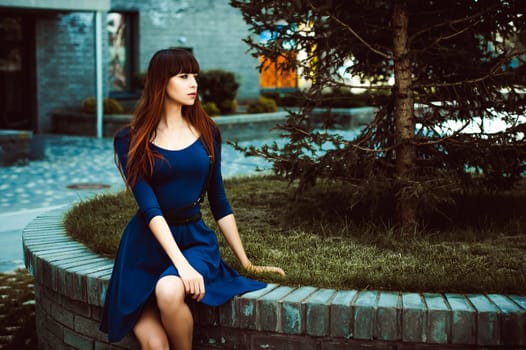 Young attractive woman with long legs with long hair in a blue elegant dress with a sword belt, strolls in a European modern courtyard, a warm summer day