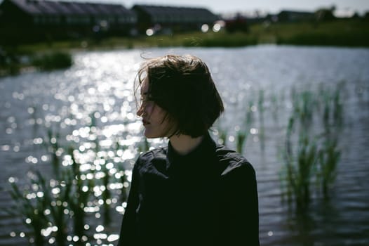 street portrait of a student girl dressed fashionably in dark clothes, against a pond background in the autumn sunny day