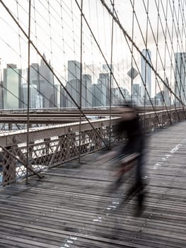Blured cyclist cycling across Brooklyn bridge. New York City Manhattan downtown skyline in sunset with skyscrapers illuminated over East River panorama as seen from Brooklyn bridge.