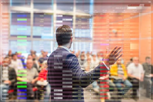 Speaker at business conference with public presentations. Audience at the conference hall. Trough the transparent presentation screen view. Charts and tables can be seen in forground.