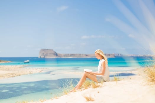 Relaxed woman enjoying sun, freedom and good book an beautiful sandy beach of Balos in Greece. Young lady reading, feeling free and relaxed. Vacations, freedom, happiness, enjoyment and well being.