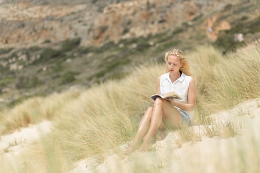 Relaxed woman enjoying sun, freedom and good book an beautiful sandy beach of Balos in Greece. Young lady reading, feeling free and relaxed. Vacations, freedom, happiness, enjoyment and well being.