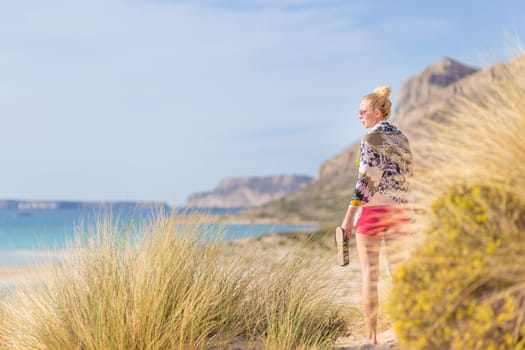 Relaxed woman wrapped in colorful scarf, enjoying sun, freedom and life at beautiful Balos beach in Greece. Concept of holidays, vacations, freedom, joy and well being.
