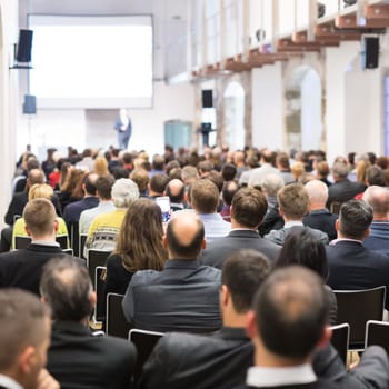Speaker giving a talk in conference hall at business event. Audience at the conference hall. Business and Entrepreneurship concept. Focus on unrecognizable businessman in the audience.