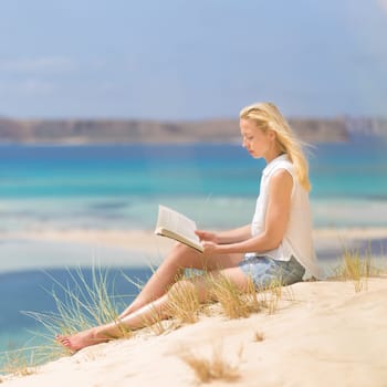 Relaxed woman enjoying sun, freedom and good book an beautiful sandy beach of Balos in Greece. Young lady reading, feeling free and relaxed. Vacations, freedom, happiness, enjoyment and well being.