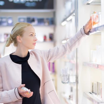 Elegant blond young woman choosing perfume in retail store. Beautiful blond lady testing and buying cosmetics in a beauty store.