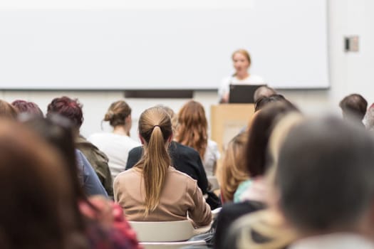 Business and entrepreneurship symposium. Female speaker giving a talk at business meeting. Audience in conference hall. Rear view of unrecognized participant in audience. Copy space on whitescreen.