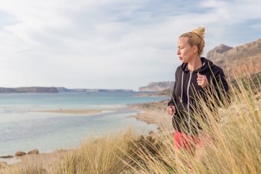 Active woman in dark hoodie running on serene Balos beach, Greece, enjoying beautiful nature and nice views. Healthy active lifestyle.